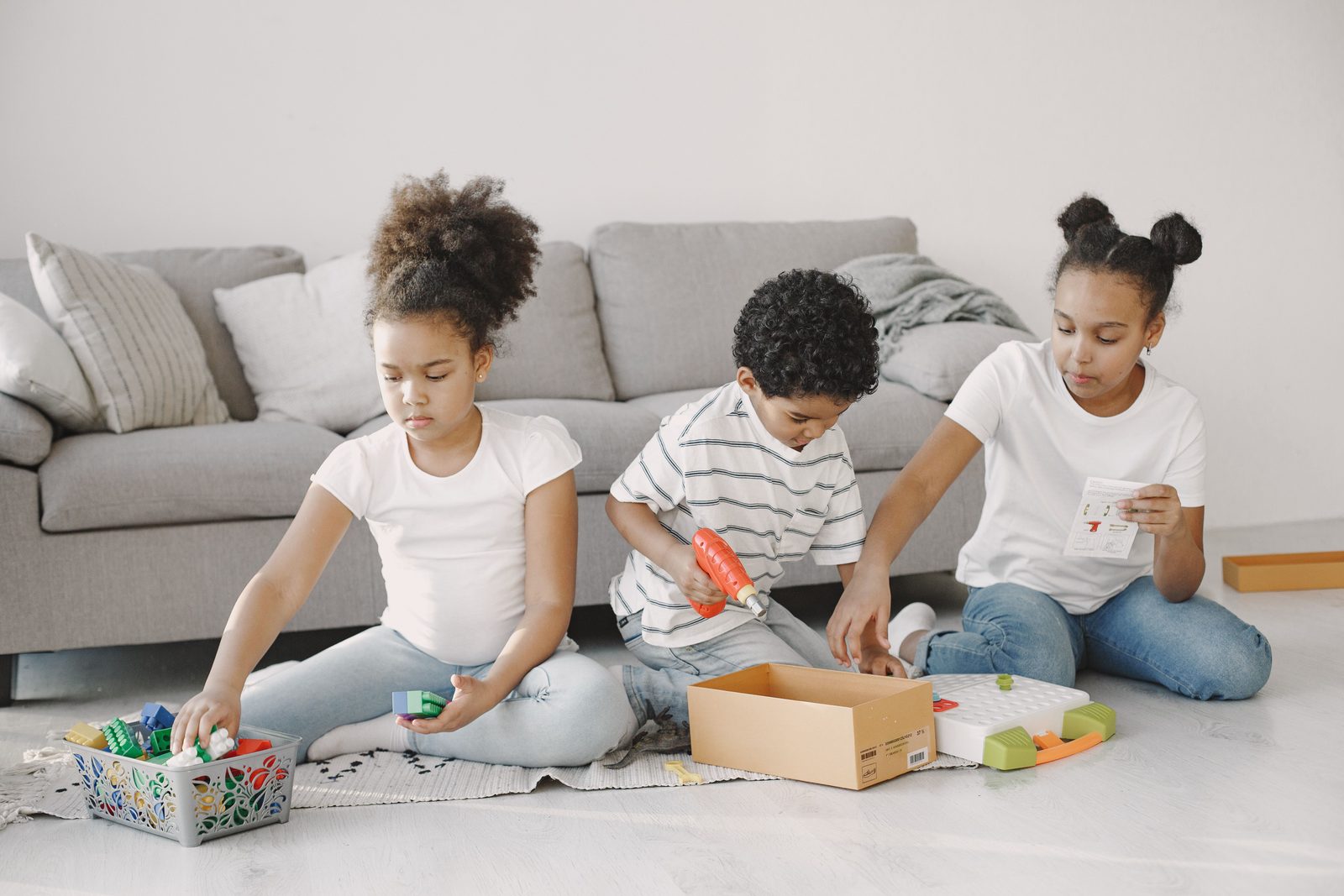 Three children sitting together on the floor of a bright living room, sorting and building with colorful blocks from a toy bin.