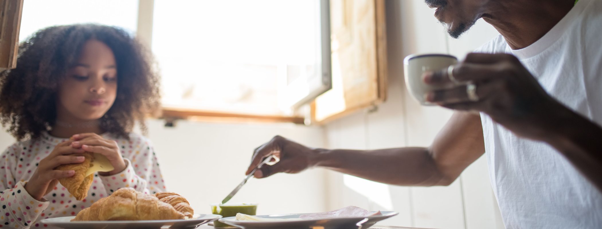 A child eating a simple home-cooked breakfast at a kitchen table beside an adult, in soft morning window light.