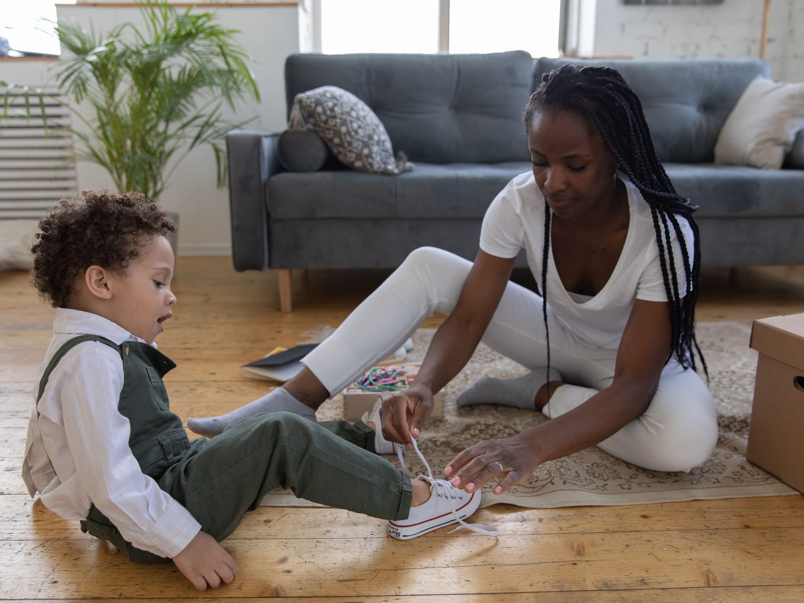 A caregiver kneeling on the living room floor, helping a young child tie his shoe.